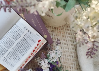 An open book with text in a non-Latin script is placed on a woven surface alongside a closed purple book. There are delicate white, purple, and light pink flowers nearby, and part of a white teapot or vase is visible.