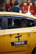 A yellow taxi with the label 'iTAKSi Kolay Taksi' is parked on a busy urban street. The car's branding includes a logo and QR code. A woman inside the taxi is smiling, while people are walking on the sidewalk behind the car. The scene suggests a typical city environment with shops and pedestrians.