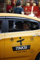 A yellow taxi with the label 'iTAKSi Kolay Taksi' is parked on a busy urban street. The car's branding includes a logo and QR code. A woman inside the taxi is smiling, while people are walking on the sidewalk behind the car. The scene suggests a typical city environment with shops and pedestrians.