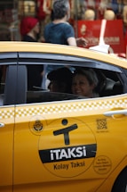 A yellow taxi with the label 'iTAKSi Kolay Taksi' is parked on a busy urban street. The car's branding includes a logo and QR code. A woman inside the taxi is smiling, while people are walking on the sidewalk behind the car. The scene suggests a typical city environment with shops and pedestrians.