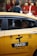 A yellow taxi with the label 'iTAKSi Kolay Taksi' is parked on a busy urban street. The car's branding includes a logo and QR code. A woman inside the taxi is smiling, while people are walking on the sidewalk behind the car. The scene suggests a typical city environment with shops and pedestrians.