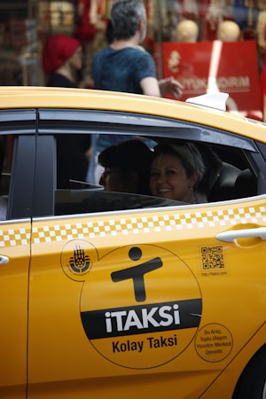 A yellow taxi with the label 'iTAKSi Kolay Taksi' is parked on a busy urban street. The car's branding includes a logo and QR code. A woman inside the taxi is smiling, while people are walking on the sidewalk behind the car. The scene suggests a typical city environment with shops and pedestrians.