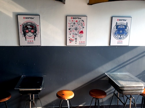 Three colorful posters featuring distinct artistic designs are mounted on a two-tone wall; the lower part is dark, while the top part is light. Below the posters, there are several stools and tables arranged against the wall.