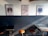 Three colorful posters featuring distinct artistic designs are mounted on a two-tone wall; the lower part is dark, while the top part is light. Below the posters, there are several stools and tables arranged against the wall.
