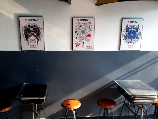 Three colorful posters featuring distinct artistic designs are mounted on a two-tone wall; the lower part is dark, while the top part is light. Below the posters, there are several stools and tables arranged against the wall.