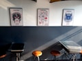 Three colorful posters featuring distinct artistic designs are mounted on a two-tone wall; the lower part is dark, while the top part is light. Below the posters, there are several stools and tables arranged against the wall.