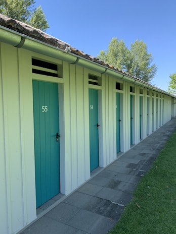 A row of light-colored beach cabins with turquoise doors, each marked with numbers. The cabins are adjacent to a paved path and grassy area, with trees visible in the background under a clear blue sky.