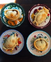 Four empanadas are placed on distinctively colored and patterned plates, each featuring floral designs. The table beneath the plates is wooden, adding a rustic touch.