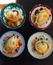 Four empanadas are placed on distinctively colored and patterned plates, each featuring floral designs. The table beneath the plates is wooden, adding a rustic touch.