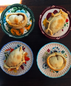 Four empanadas are placed on distinctively colored and patterned plates, each featuring floral designs. The table beneath the plates is wooden, adding a rustic touch.