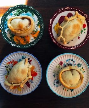 Four empanadas are placed on distinctively colored and patterned plates, each featuring floral designs. The table beneath the plates is wooden, adding a rustic touch.
