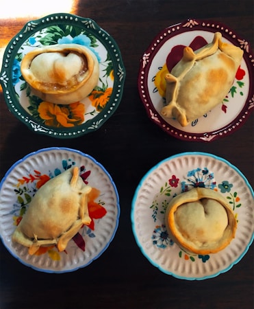 Four empanadas are placed on distinctively colored and patterned plates, each featuring floral designs. The table beneath the plates is wooden, adding a rustic touch.