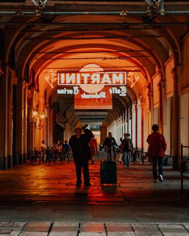 An urban scene depicting a covered walkway with people walking and sitting at tables. A man stands in the center holding a travel bag with a neon sign above casting an orange glow.