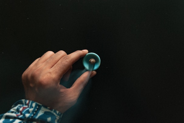 A hand is holding a spinning turquoise top against a dark background. The person is wearing a long-sleeved garment with a colorful, geometric pattern. The lighting creates a subtle shadow, adding depth to the image.
