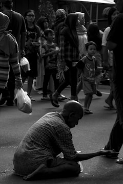 A black and white image depicts a busy street scene. An elderly person is sitting on the ground with an outstretched arm, possibly asking for help or money. Surrounding them, many people are walking by, seemingly engaged in their activities. Some are carrying shopping bags, and children are present in the crowd.
