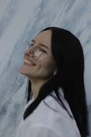 A person with long dark hair and glasses smiles broadly, showing dimples, against a textured background that resembles marble.