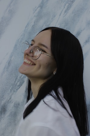 A person with long dark hair and glasses smiles broadly, showing dimples, against a textured background that resembles marble.