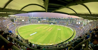A large cricket stadium filled with spectators. The field is bright green, and players are positioned in the center. Multiple levels of tiered seating are visible, with a covered roof structure above. The stadium is illuminated with lights, and the city skyline is visible in the background.