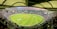 A large cricket stadium filled with spectators. The field is bright green, and players are positioned in the center. Multiple levels of tiered seating are visible, with a covered roof structure above. The stadium is illuminated with lights, and the city skyline is visible in the background.