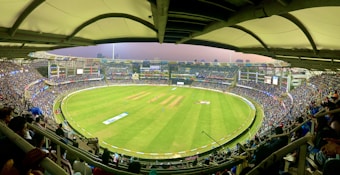 A large cricket stadium filled with spectators. The field is bright green, and players are positioned in the center. Multiple levels of tiered seating are visible, with a covered roof structure above. The stadium is illuminated with lights, and the city skyline is visible in the background.