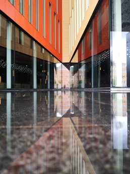 The image features a modern architectural structure with reflective glass surfaces. The lines and geometric shapes create a symmetrical pattern as they converge towards a vanishing point. The reflection on the floor enhances the sense of symmetry and depth. The buildings are composed of red, beige, and brown tones, with a mix of vertical and horizontal lines.