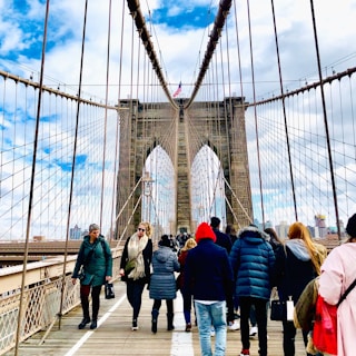 A group of people walking across the iconic structure of the Brooklyn Bridge. The suspension cables create a geometric pattern against a partly cloudy sky. The skyline of New York City is faintly visible in the background.
