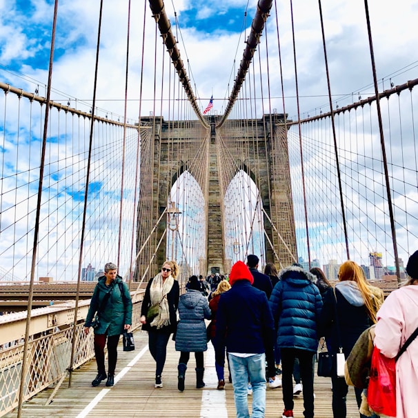A group of people walking across the iconic structure of the Brooklyn Bridge. The suspension cables create a geometric pattern against a partly cloudy sky. The skyline of New York City is faintly visible in the background.