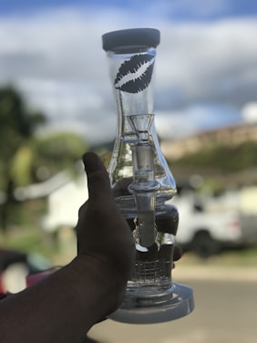 A hand holds a clear glass object with a round base and a cylindrical top featuring a black lip print design. The background is a suburban neighborhood with blurred greenery and vehicles, suggesting a casual outdoor setting.
