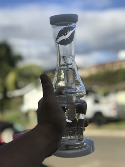 A hand holds a clear glass object with a round base and a cylindrical top featuring a black lip print design. The background is a suburban neighborhood with blurred greenery and vehicles, suggesting a casual outdoor setting.