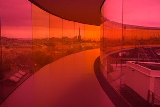 A panoramic walkway enclosed by vibrant red and orange glass panels provides a tinted view of an urban skyline. The ceiling is curved, and the floor follows a smooth path, enhancing the futuristic atmosphere. The view includes diverse architecture from the city, with buildings and a prominent spire visible in the distance.