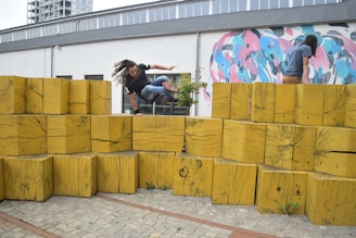A man is performing parkour over large, stacked wooden blocks outside a building with a colorful mural on the wall. Another person is on top of the blocks, preparing to jump or climb.