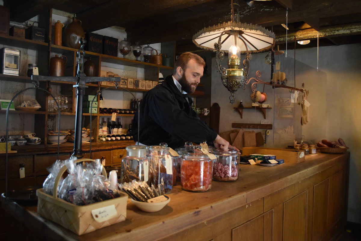 Un homme barbu en veste noire range des bocaux de bonbons sur un long comptoir en bois, devant des étagères garnies et sous une lampe ancienne