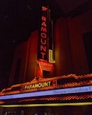 A vintage-style theater marquee with the word 'Paramount' illuminated in bright red and yellow lights. The vertical and horizontal sign structures are adorned with detailed neon lighting against a dark background. Below, a digital marquee displays part of a message about Emerson College.