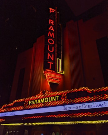 A vintage-style theater marquee with the word 'Paramount' illuminated in bright red and yellow lights. The vertical and horizontal sign structures are adorned with detailed neon lighting against a dark background. Below, a digital marquee displays part of a message about Emerson College.