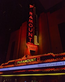 A vintage-style theater marquee with the word 'Paramount' illuminated in bright red and yellow lights. The vertical and horizontal sign structures are adorned with detailed neon lighting against a dark background. Below, a digital marquee displays part of a message about Emerson College.