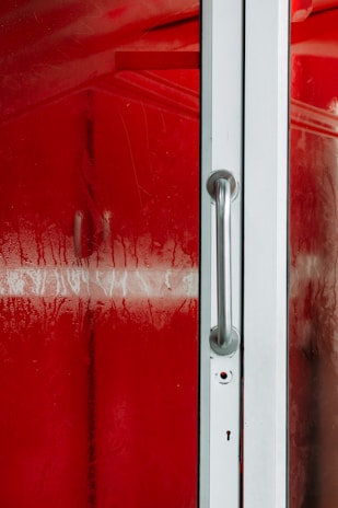 A close-up view of a glass door with a metallic handle on a red, foggy background. The surface of the glass is moist, with visible condensation and droplets.