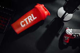 A vibrant red shaker with 'CTRL A Meal Replacement' printed on it rests on a dark surface, accompanied by a gaming controller with red accents and a black microphone on a stand. Part of a keyboard is visible on the left, partially illuminated by red backlight.