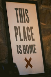 A framed sign leaning against a textured stone wall. The sign has a bold message in black capital letters saying 'THIS PLACE IS HOME', with a large brown cross below the text.