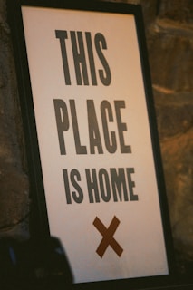 A framed sign leaning against a textured stone wall. The sign has a bold message in black capital letters saying 'THIS PLACE IS HOME', with a large brown cross below the text.