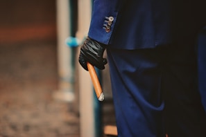 A person dressed in a dark blue suit and wearing black gloves holds a large cigar. The focus is on the details of the suit buttons and the cigar.