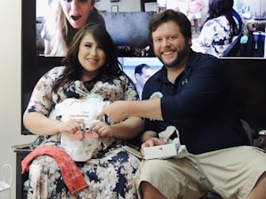 A happy couple is sitting together, displaying a baby onesie and small shoes. The woman is wearing a floral dress, while the man is in a dark shirt and khaki shorts. Behind them is a screen showing people on a video call, likely participating in the event remotely. Various small items and decorations are in the background.