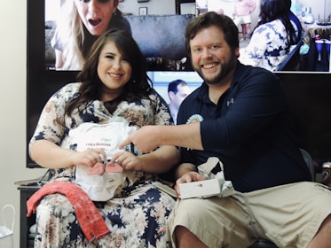 A happy couple is sitting together, displaying a baby onesie and small shoes. The woman is wearing a floral dress, while the man is in a dark shirt and khaki shorts. Behind them is a screen showing people on a video call, likely participating in the event remotely. Various small items and decorations are in the background.