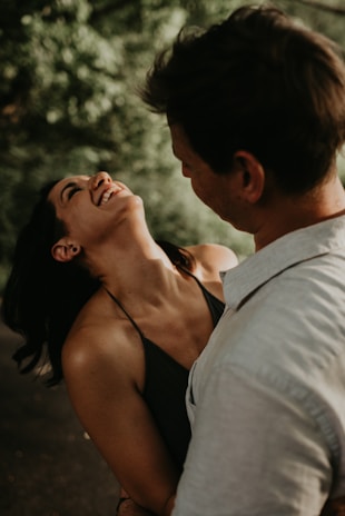 A couple is captured in a joyful and intimate moment, with the woman laughing heartily and leaning back in a happy pose while the man looks at her warmly. The background is lush and green, suggesting a setting surrounded by nature.