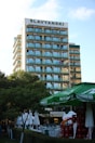 A multi-story hotel building with a sign labeled SLAVYANSKI at the top. The building has multiple balconies with glass railings. In the foreground, there are green trees, large green umbrellas, and stacks of red crates. There are also white outdoor umbrellas and tables set up near a hedge.