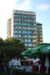 A multi-story hotel building with a sign labeled SLAVYANSKI at the top. The building has multiple balconies with glass railings. In the foreground, there are green trees, large green umbrellas, and stacks of red crates. There are also white outdoor umbrellas and tables set up near a hedge.