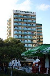 A multi-story hotel building with a sign labeled SLAVYANSKI at the top. The building has multiple balconies with glass railings. In the foreground, there are green trees, large green umbrellas, and stacks of red crates. There are also white outdoor umbrellas and tables set up near a hedge.