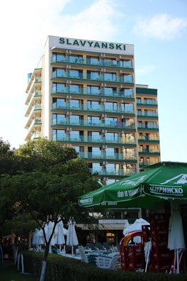 A multi-story hotel building with a sign labeled SLAVYANSKI at the top. The building has multiple balconies with glass railings. In the foreground, there are green trees, large green umbrellas, and stacks of red crates. There are also white outdoor umbrellas and tables set up near a hedge.