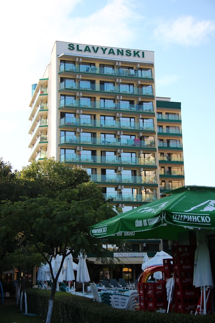 A multi-story hotel building with a sign labeled SLAVYANSKI at the top. The building has multiple balconies with glass railings. In the foreground, there are green trees, large green umbrellas, and stacks of red crates. There are also white outdoor umbrellas and tables set up near a hedge.