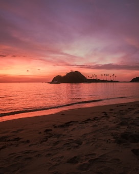 A serene beach scene during sunset with a silhouette of an island in the distance. The sky is painted with vibrant hues of orange, pink, and purple, reflecting off the calm ocean waters. Palm trees are visible on the island, adding to the tropical feel.
