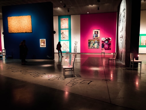 An art gallery with colorful illuminated walls displaying various paintings and artworks. Dimly lit space with modern and abstract art, featuring multiple framed pieces including a large monochrome portrait. Chairs are strategically placed along the walls, and subtle lighting is used to highlight specific artworks.
